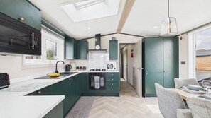 A compact kitchen with a microwave, stove, and sink, a dining area with chairs, and a view through a door. - Bumblebee - 2 Bedroom Lodge - Manorbier (Tenby)