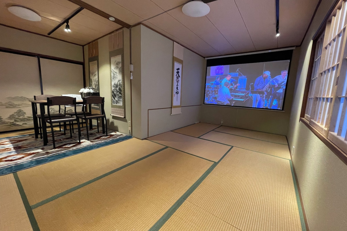 Een traditionele kamer met tatami matten, met een televisiescherm, een tafel met stoelen en een decoratieve wand.