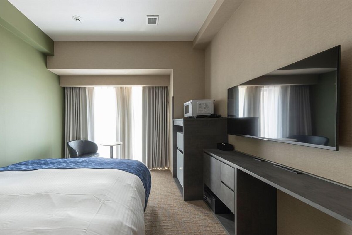 A hotel room with a bed, a desk with a TV, a chair, and a window with curtains.