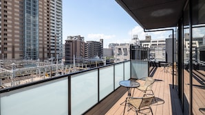 A balcony with a glass railing overlooking a cityscape with buildings and a train track. There are two wicker chairs and a small round table. - Hotel R9 Premium Ichikawa Ekimae (Ichikawa)