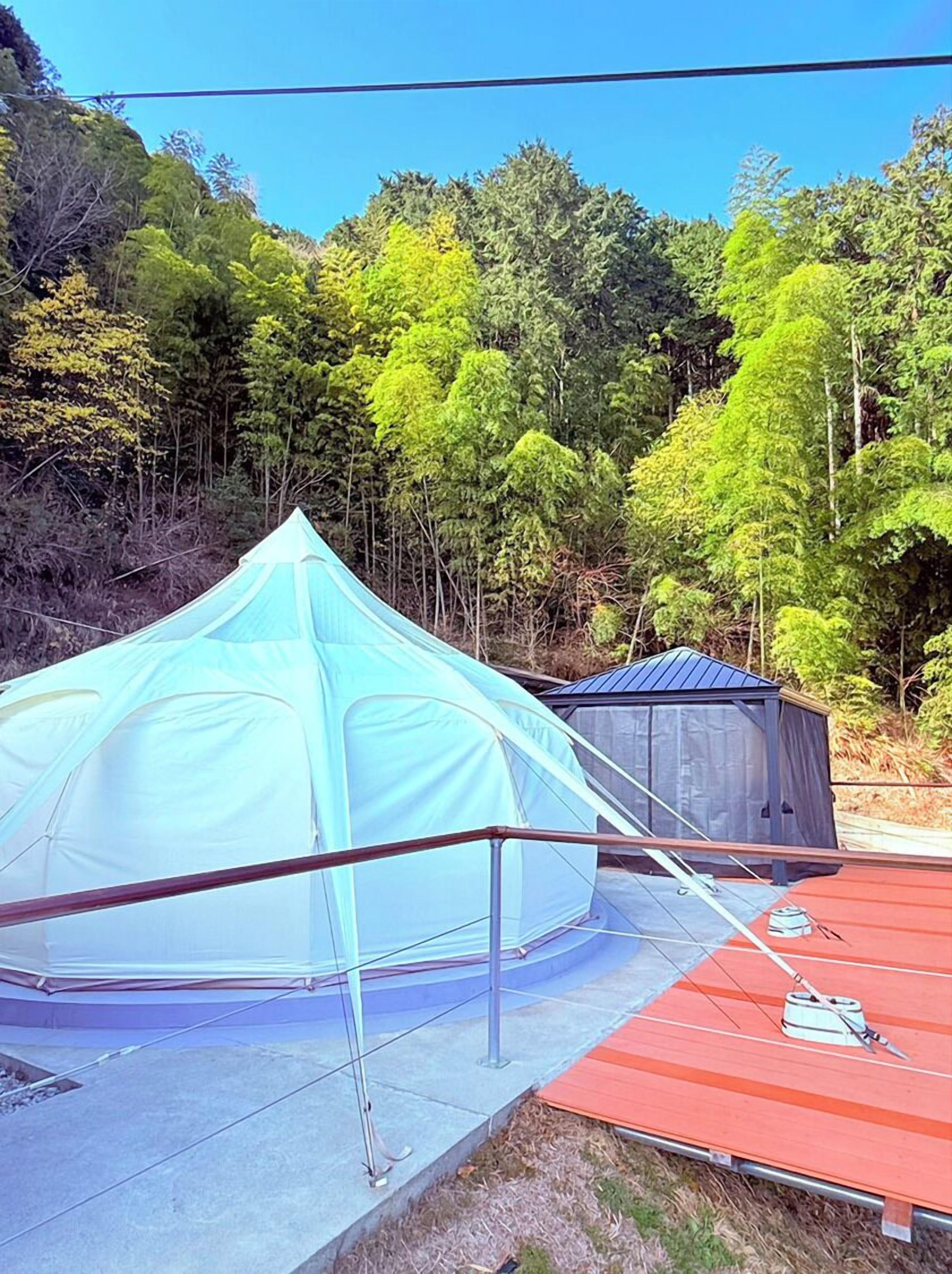 A tent with a metal frame and a smaller structure nearby, set against a backdrop of trees.
