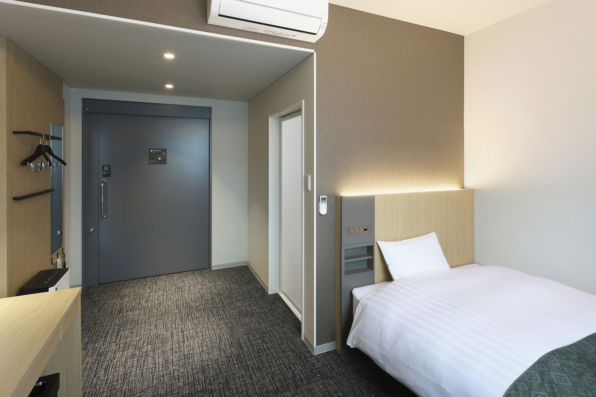 A hotel room with a bed, a desk, a chair, a wardrobe, and a door.