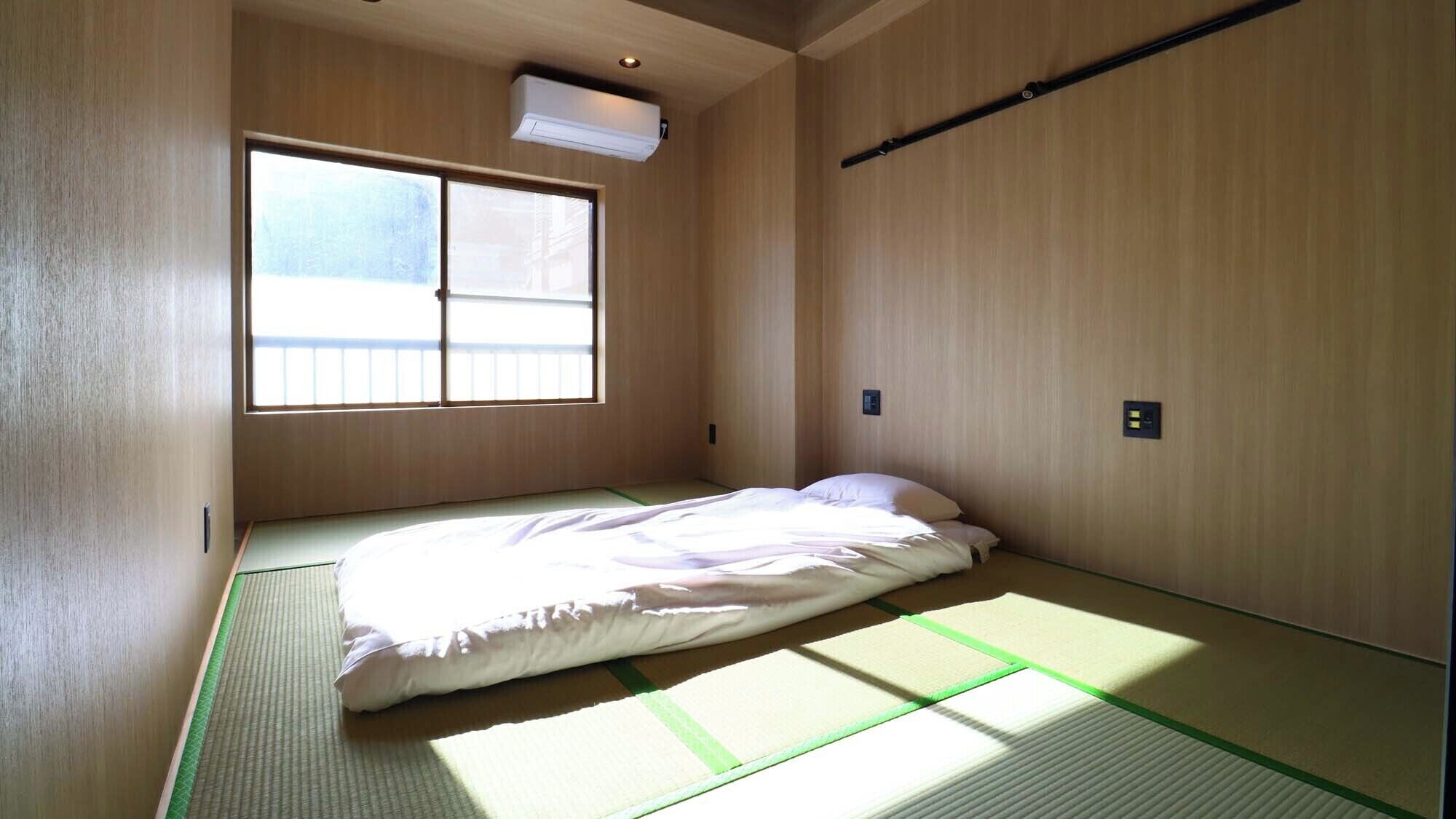 A small, minimalist room with tatami flooring, a single bed, a window, and a ceiling-mounted air conditioning unit.