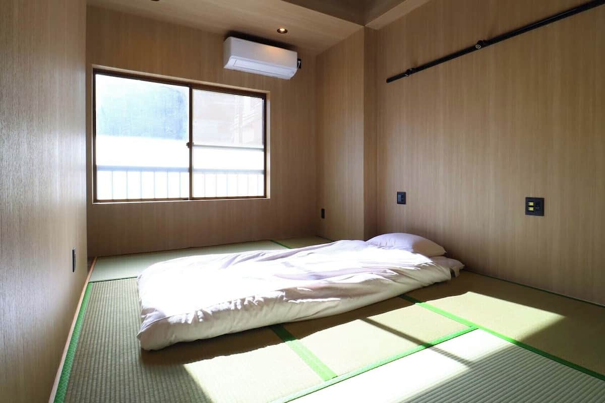 A small, minimalist room with tatami flooring, a single bed, a window, and a ceiling-mounted air conditioning unit.