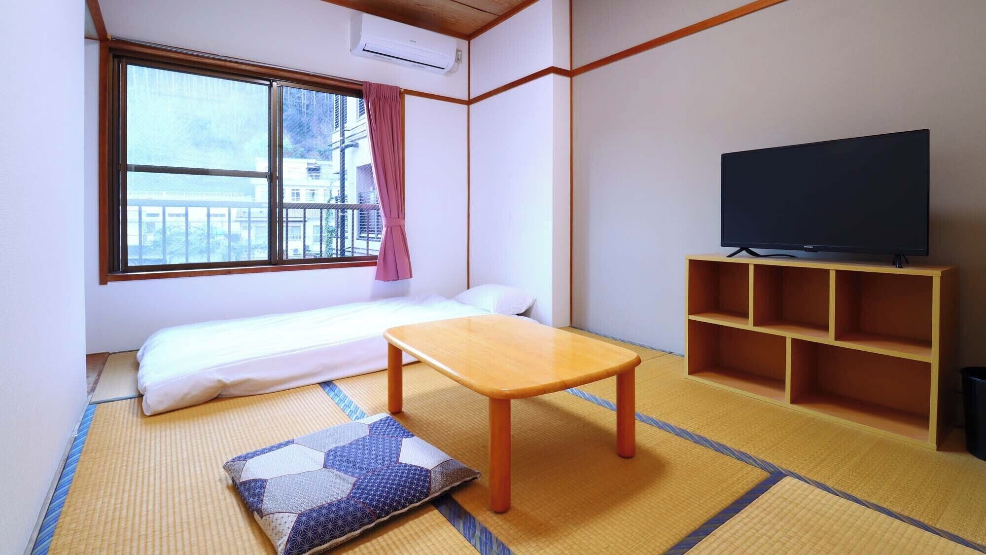 A room with a sofa bed, a television on a wooden stand, tatami mats, and a window with a view of buildings.