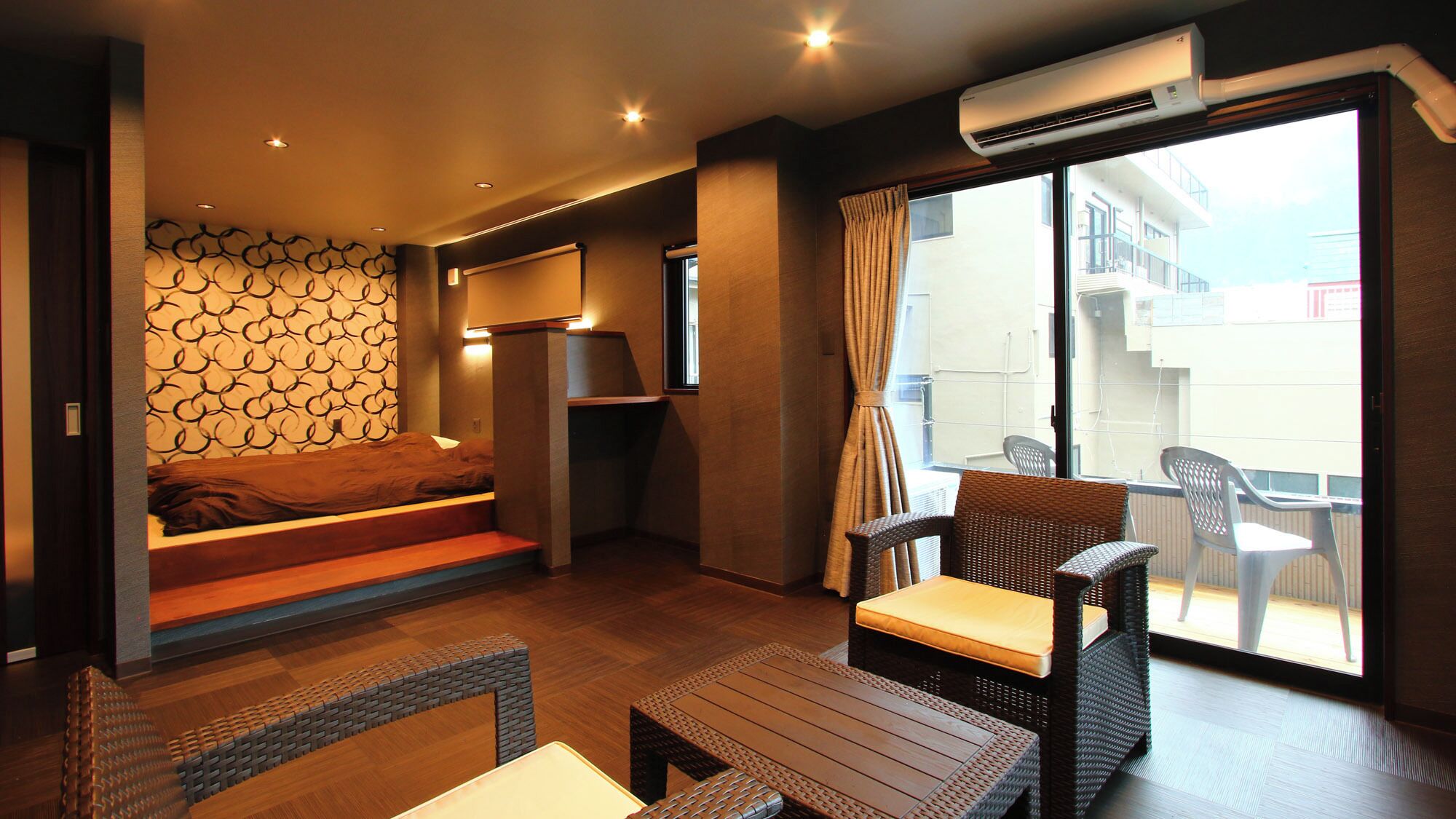 A compact hotel room with a bed, a small table, wicker chairs, a balcony with plastic chairs, and a patterned wall.
