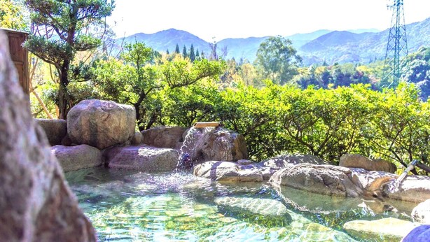A clear outdoor water feature with rocks and a bamboo structure, surrounded by lush greenery and mountains in the background. - Kumanokawa Onsen Minshuku Mimi (Saga)