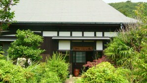 A traditional Japanese building with a sloped roof, surrounded by lush greenery and a small pathway leading to the entrance. - Hananoki Inn (Sado)