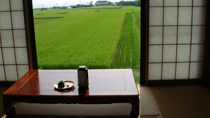 A room with tatami flooring, a wooden table with a tea set, and a view of a green field through shoji screens. - Hananoki Inn (Sado)
