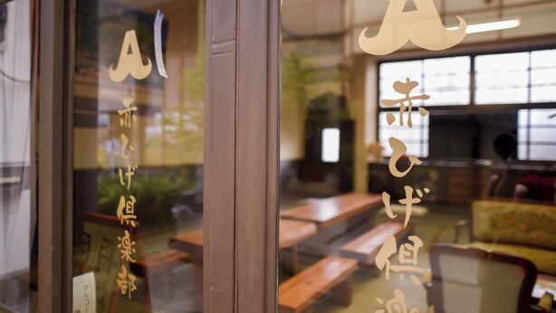 Interior de un restaurante con mesas y sillas de madera, y letras grandes en la puerta de vidrio.