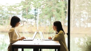 Dos personas trabajando en una mesa con computadoras portátiles, cerca de una ventana que da a vista de árboles y una masa de agua.
