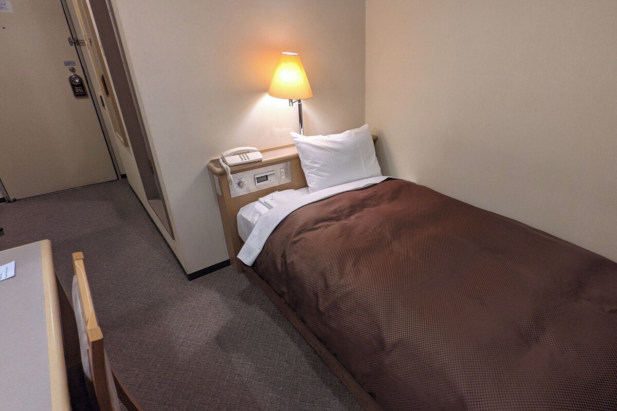 A hotel room with a single bed, a bedside table with a lamp, a phone, and a small desk with a chair.