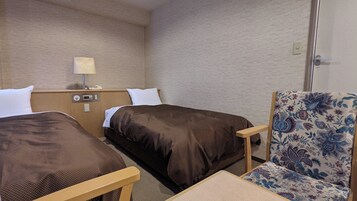 Habitación de hotel con dos camas individuales, un escritorio pequeño con lámpara y un sillón con estampado floral.