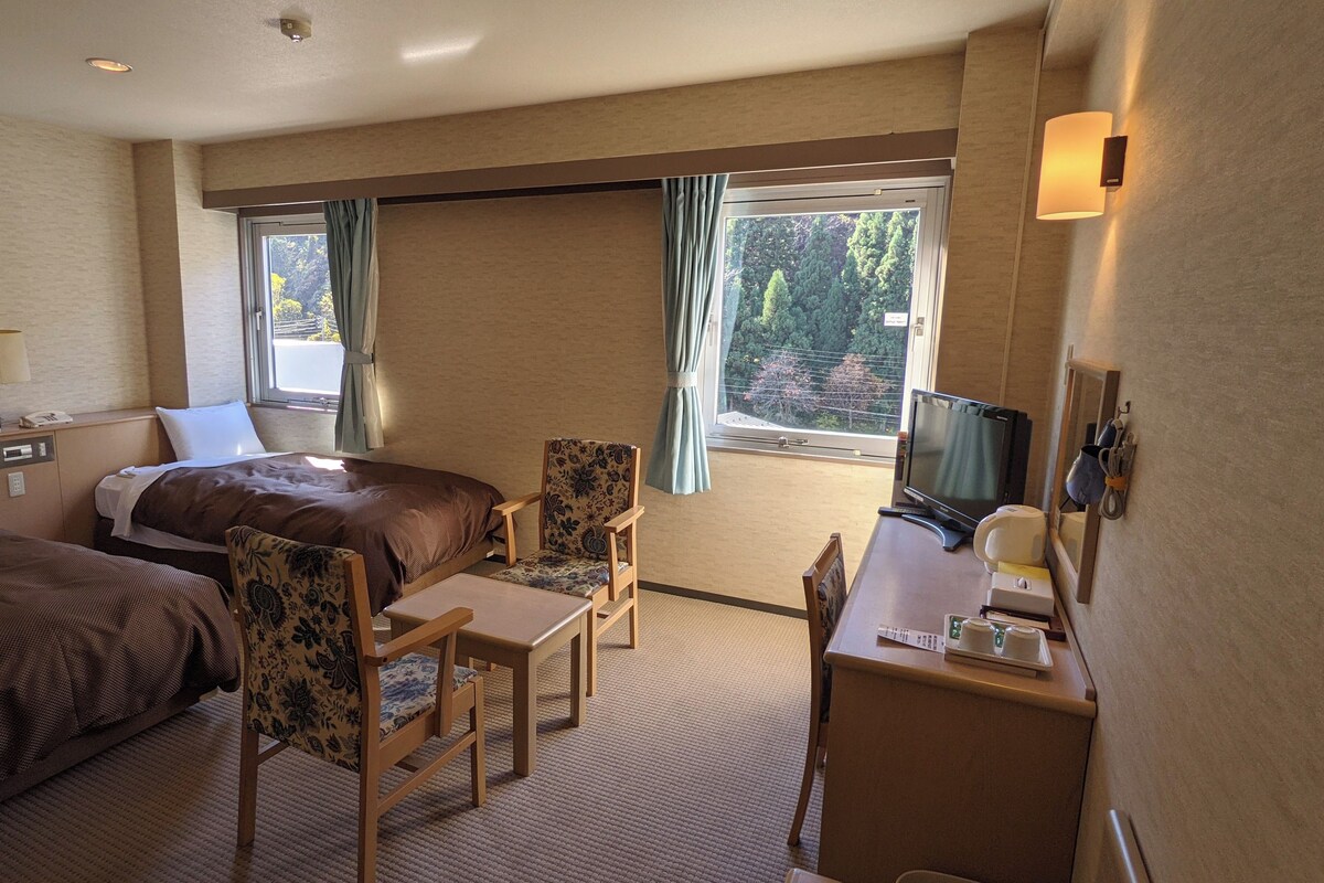 A hotel room with a bed, a desk with a computer, a chair, a TV, and a window with curtains.