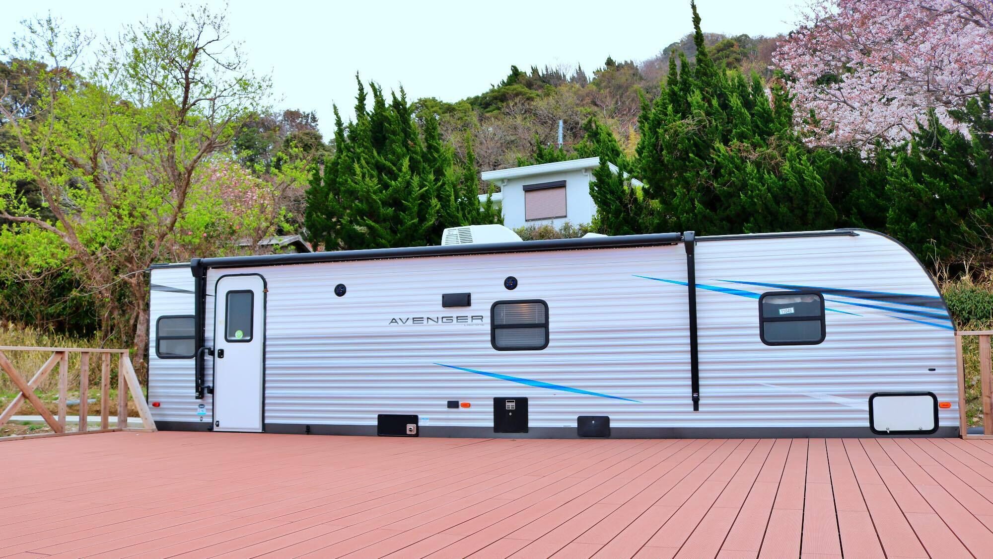 A stationary RV with a white exterior and blue stripes, parked on a wooden deck with a backdrop of trees and a hill.