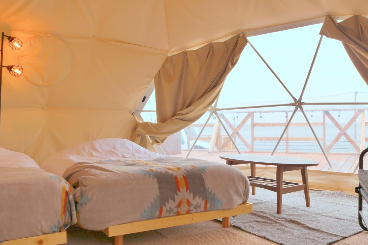 A cozy tented accommodation with two beds, a small table, and a view of the sea.