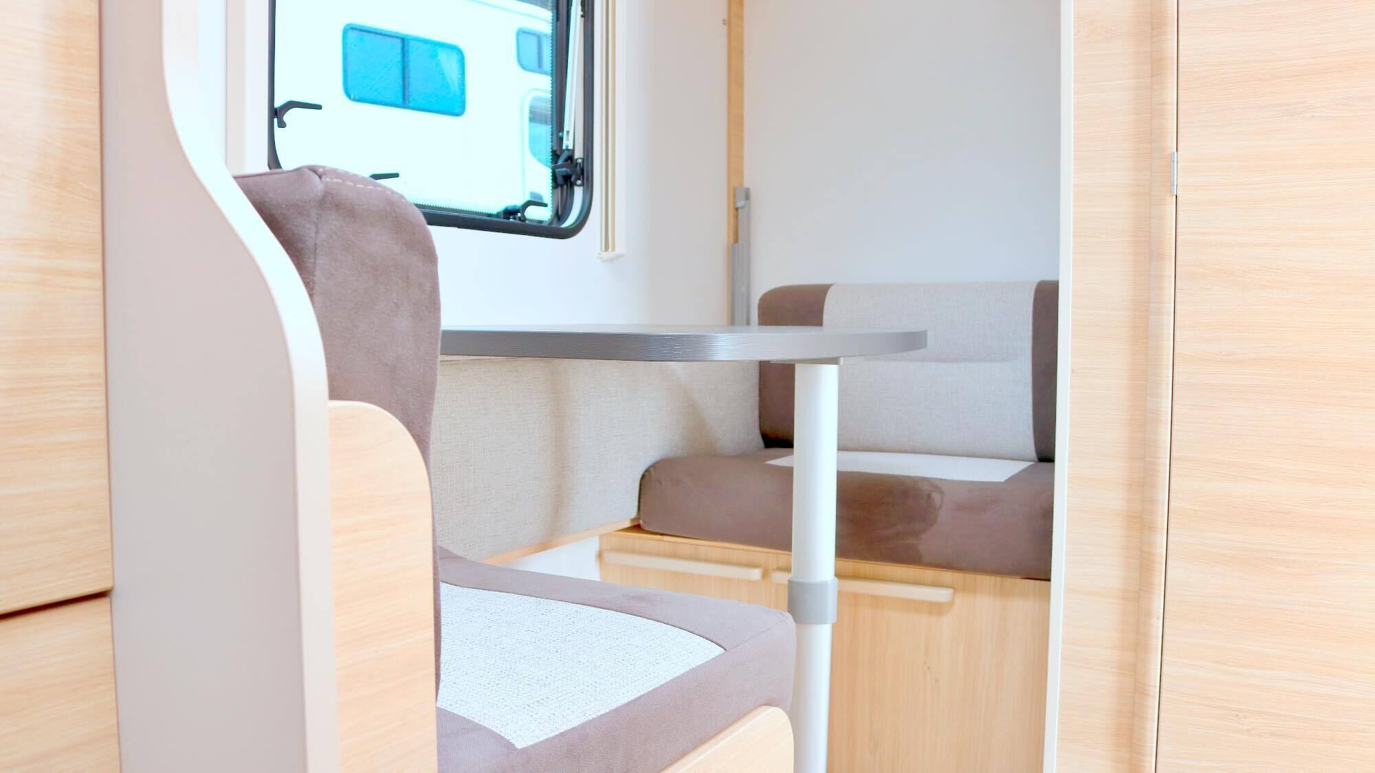 A compact RV interior with a dining area, a window with blinds, and a kitchenette.