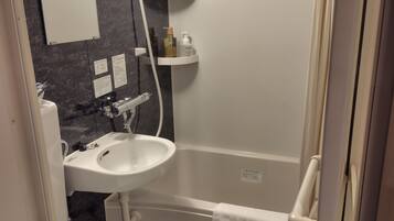 Un baño con lavamanos, inodoro y bañera blancos. Hay estantes con artículos de aseo y una toalla colgada en la pared.