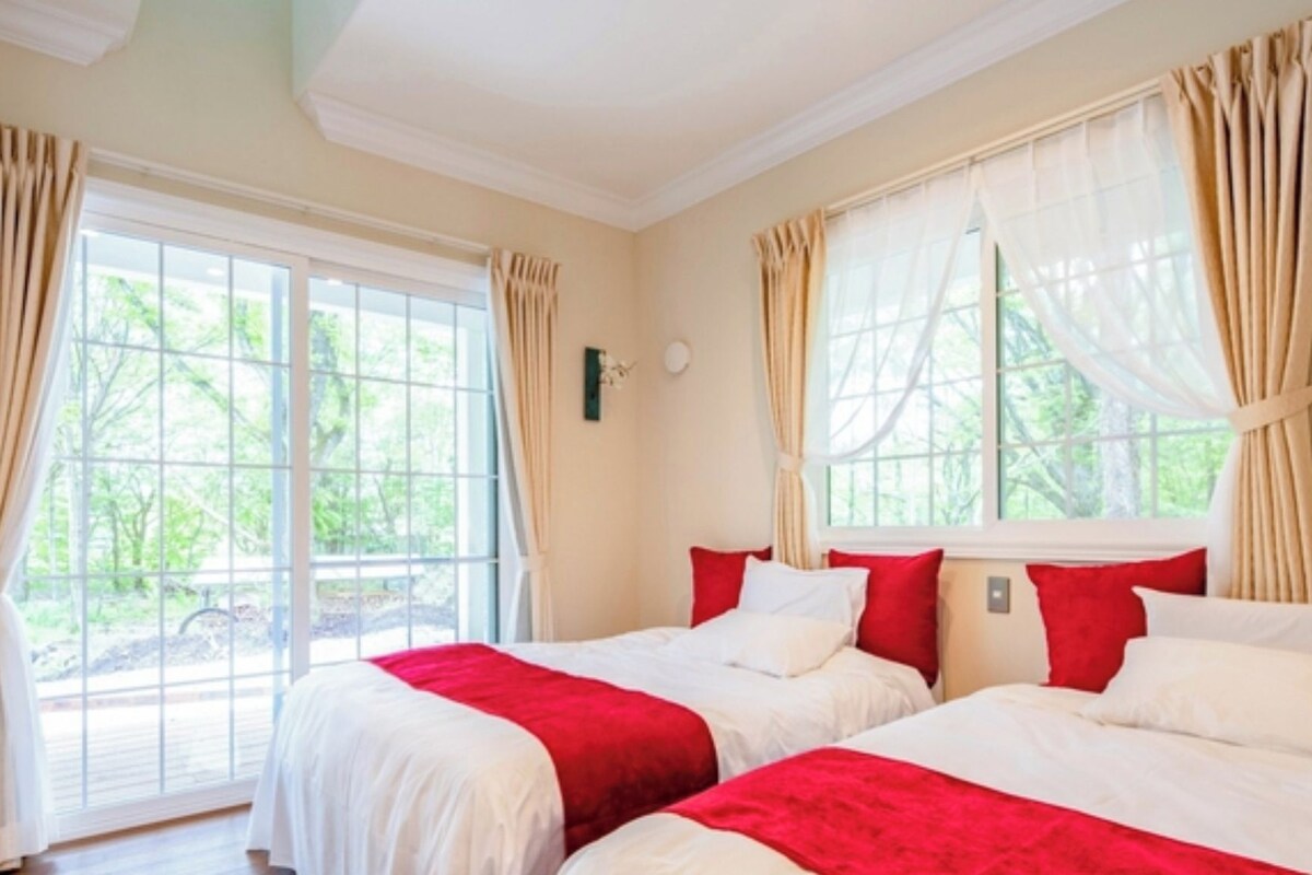 A hotel room with two beds, beige curtains, and large windows offering a view of greenery.
