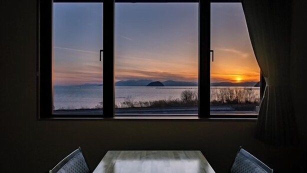 Una habitación con un gran ventanal que da a una puesta de sol sobre el agua, con una mesa y dos sillas al frente.