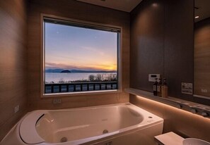 A modern bathroom with a large bathtub, a window with a view of a sunset over water, and a shelf with toiletries. - Ryoshi no Yado Funaso (Nagahama)