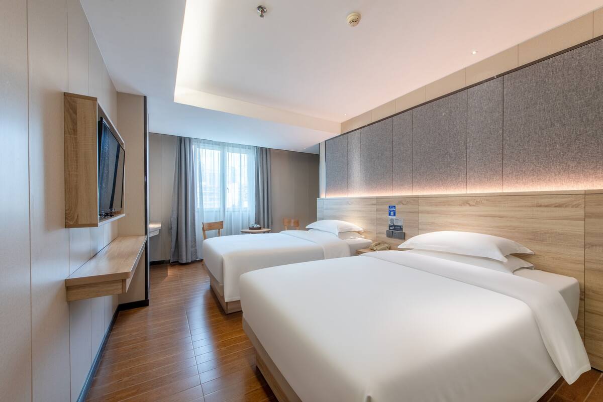 A hotel room with two beds, a TV mounted on the wall, a bedside table, a lamp, and a window with curtains.