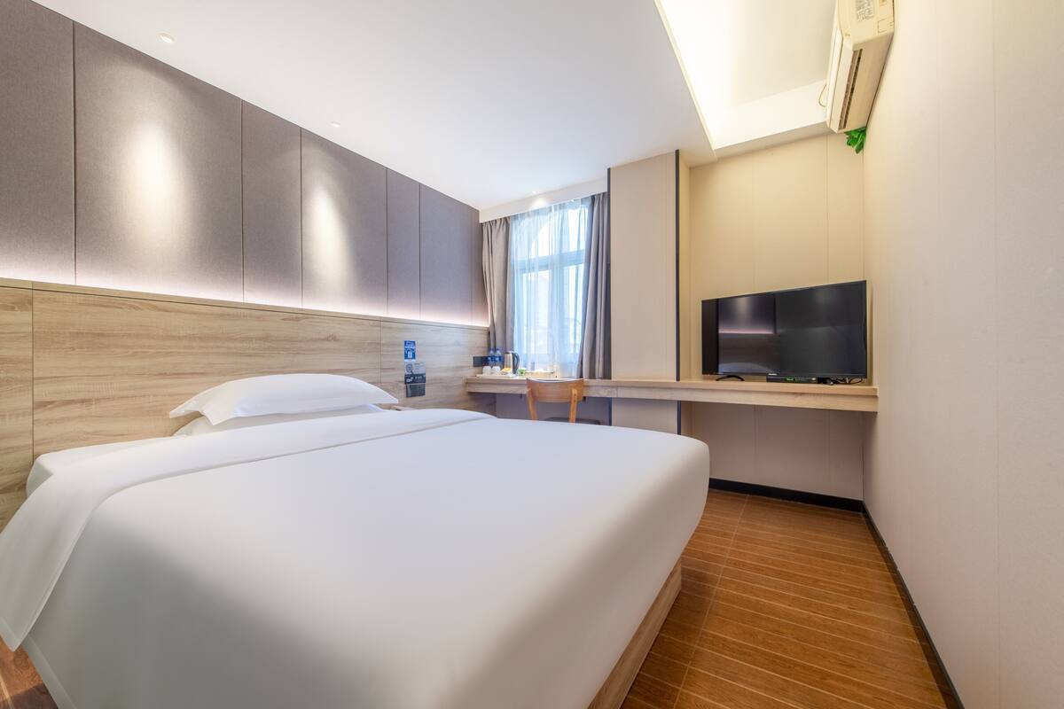 A hotel room with a large bed, a desk with a chair, a television, and an air conditioning unit.