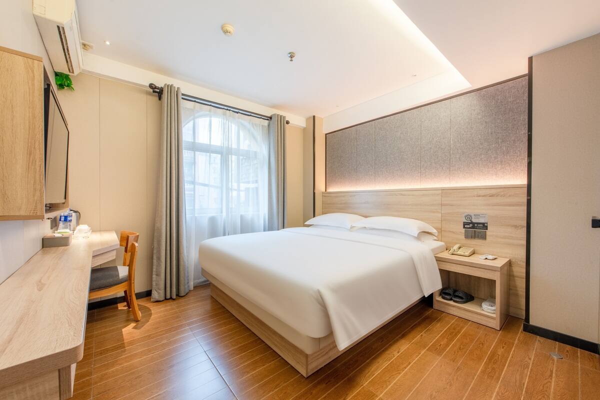A modern hotel room with a wooden floor, a bed with white linens, a desk with a chair, a nightstand with a phone and shoes, and a large window with curtains.