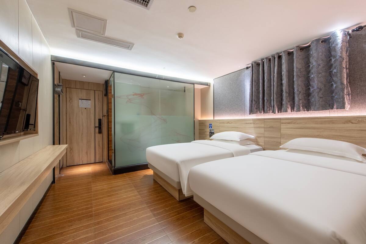 A hotel room with two beds, a wooden floor, a glass door leading to a bathroom, and a mounted TV.