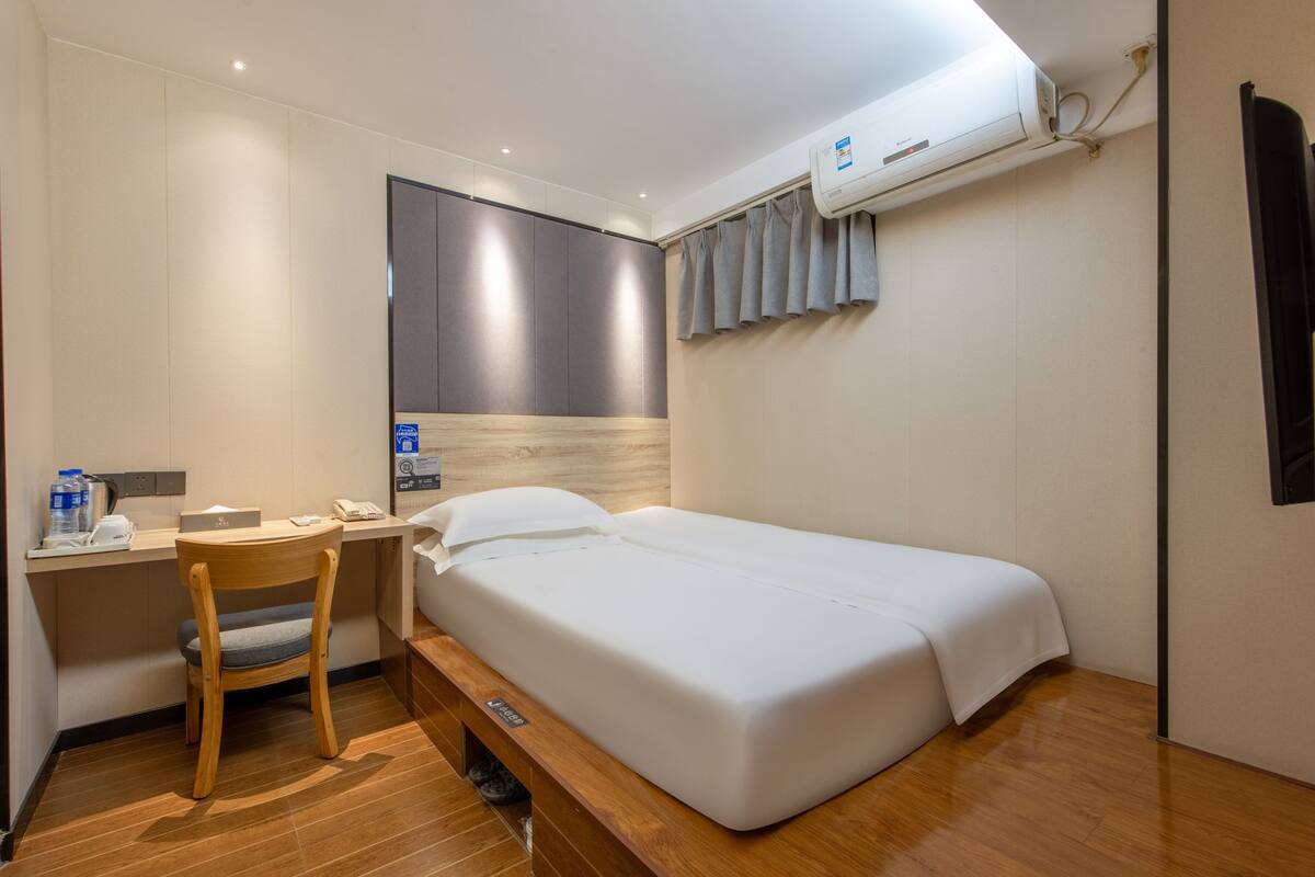 A hotel room with a bed, a desk with a chair, a telephone, and an air conditioning unit.