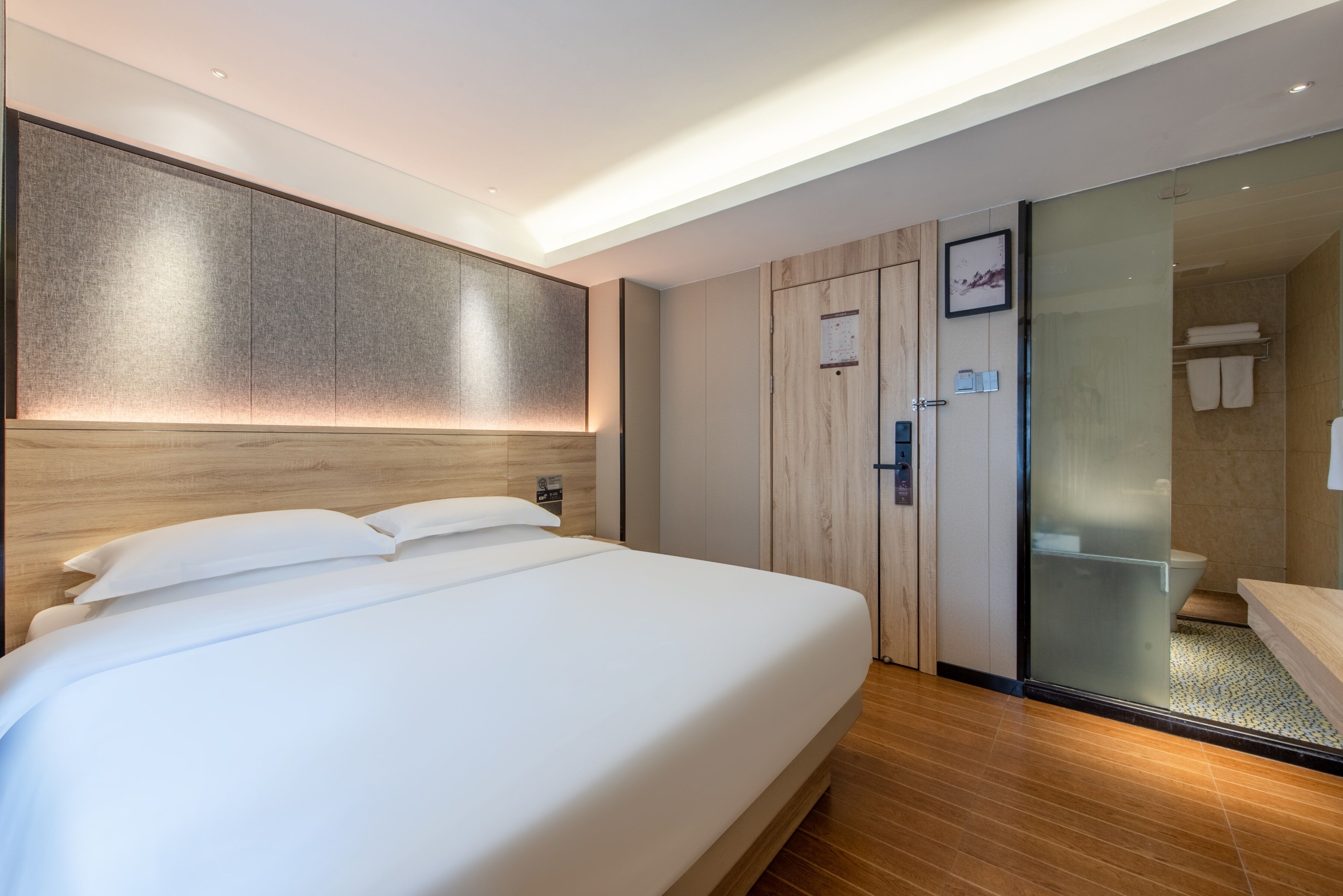 Ein modernes Hotelzimmer mit einem großen Bett, einem Holz-Kopfteil, einer Schiebetür zur Badezimmereinrichtung und einem Holzboden.