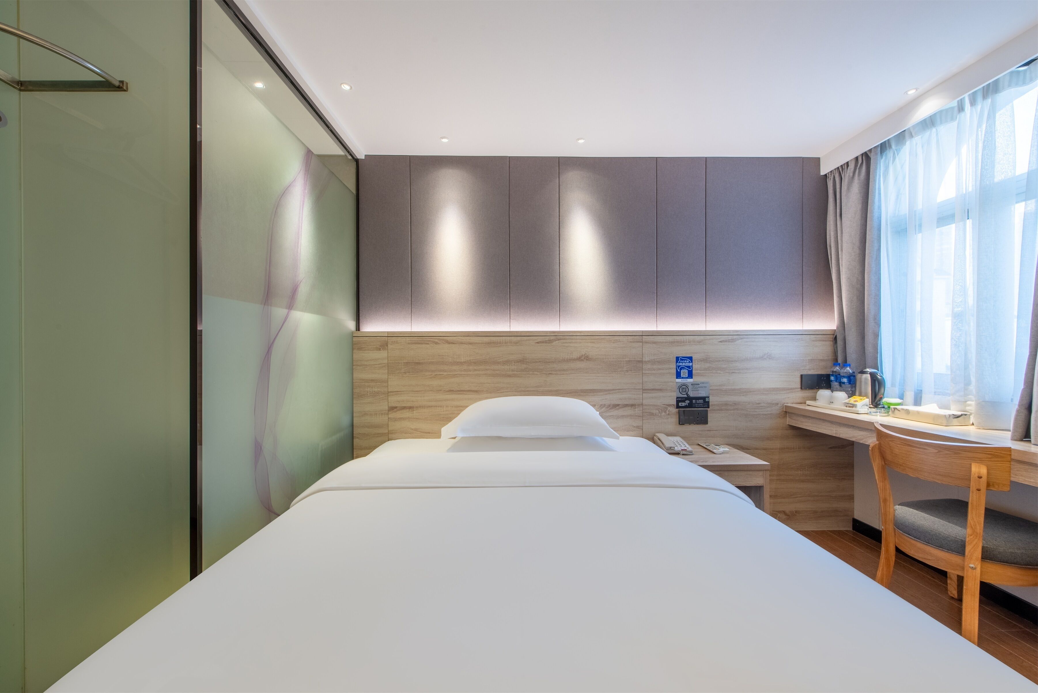 A modern hotel room with a large bed, a desk with a chair, a glass partition, and a wooden headboard.