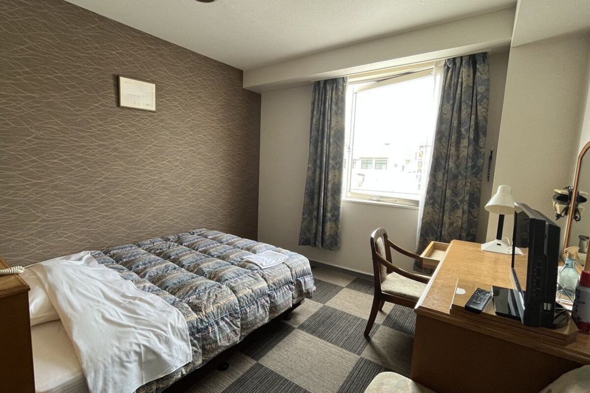 A hotel room with a bed, a desk, a chair, a lamp, a telephone, and a window with curtains.