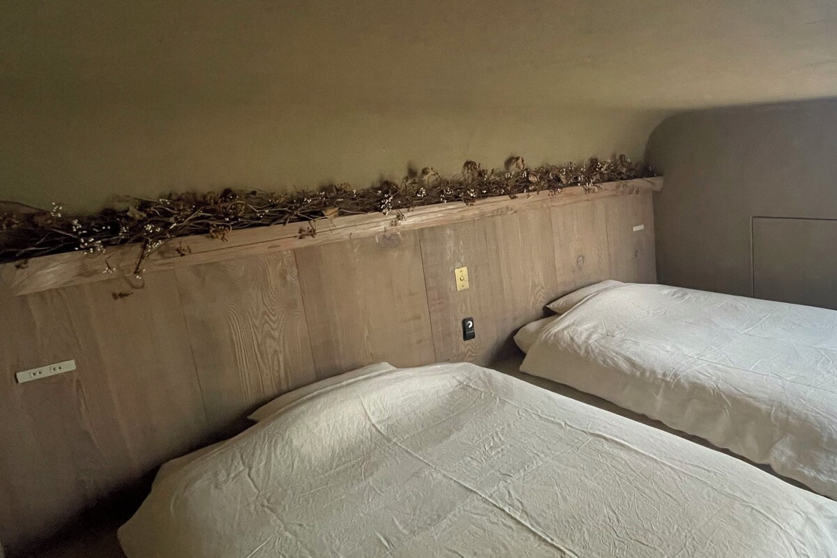 A room with two beds, a wooden headboard, a wall-mounted light switch, and a door.