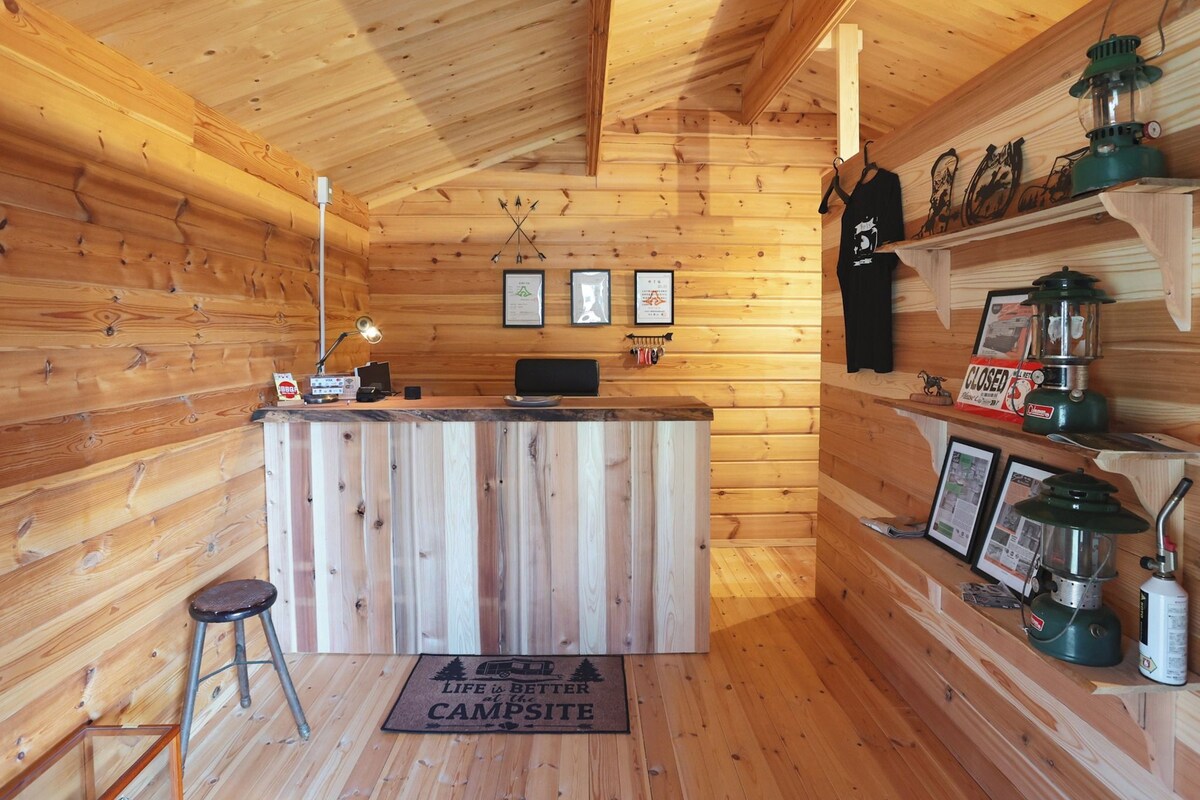 Interieur van een houten hutje met een receptiebalie, een bureaulamp, ingelijste foto's, een zwart schort en kampeergerelateerde spullen op planken.