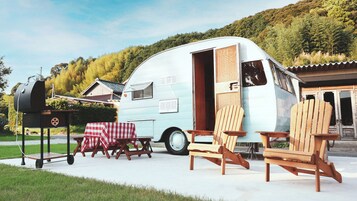 Una caravana antigua con una puerta corredera, una parrilla para barbacoa y dos sillas Adirondack.