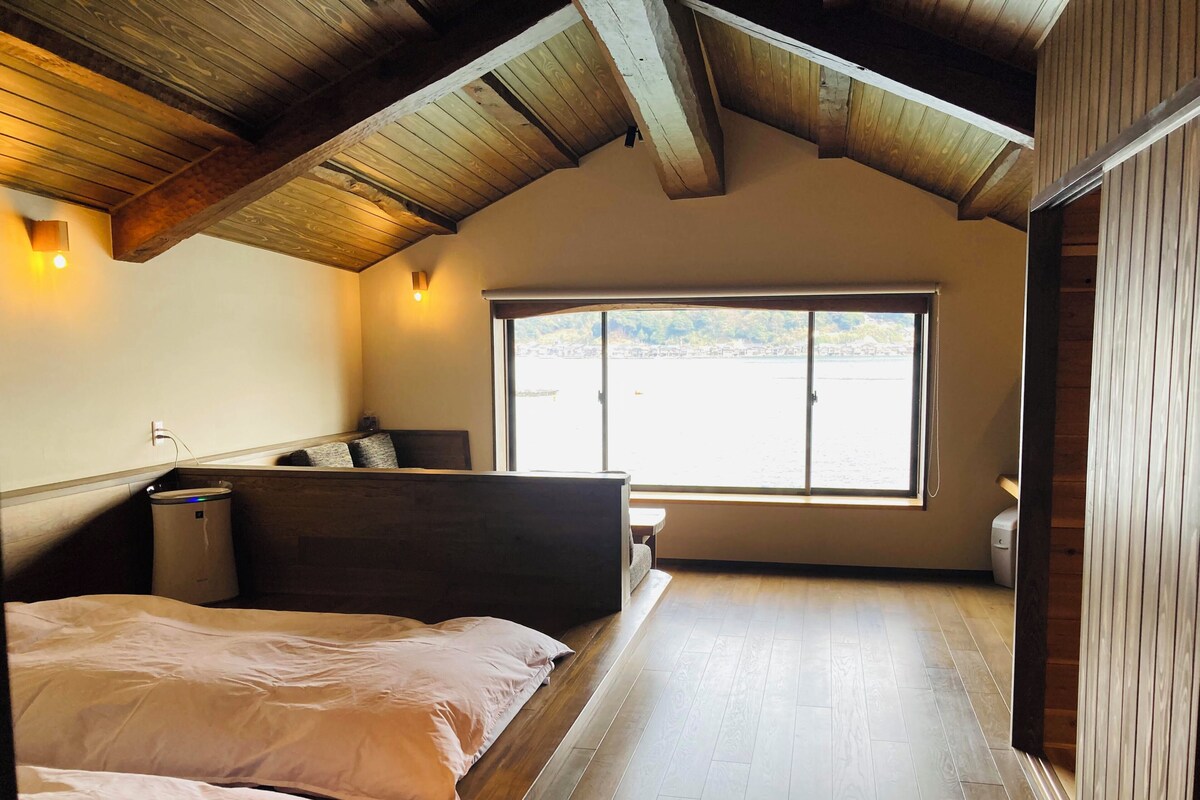 A bedroom with a wooden ceiling, a large window, a bed, and a bedside table.