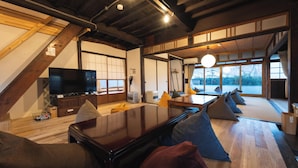 A modern interior with a wooden dining table, cushioned seating, a flat-screen TV, and large windows offering a view of the outdoors. - Teihaku Chichibu Nagatoro Folk House Rental (Nagatoro)