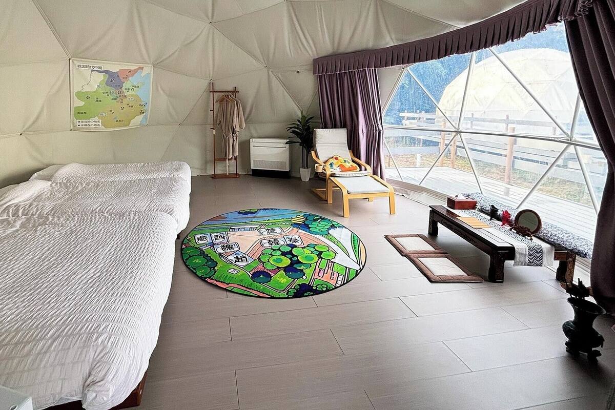 A tented room with a bed, a round rug, a chair, a table, and a map on the wall.