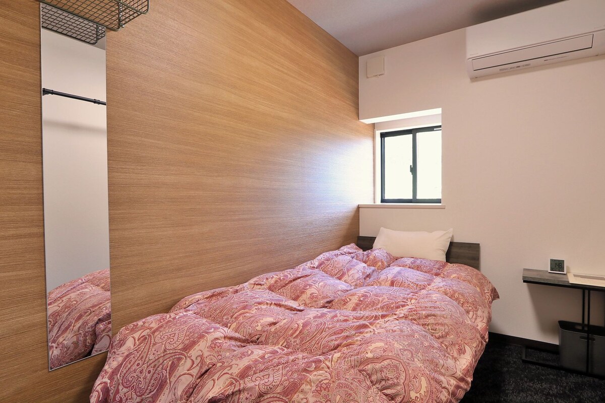 A modern bedroom with a large mirror, a bed with a patterned duvet, a bedside table, a small nightstand, and a window with natural light.