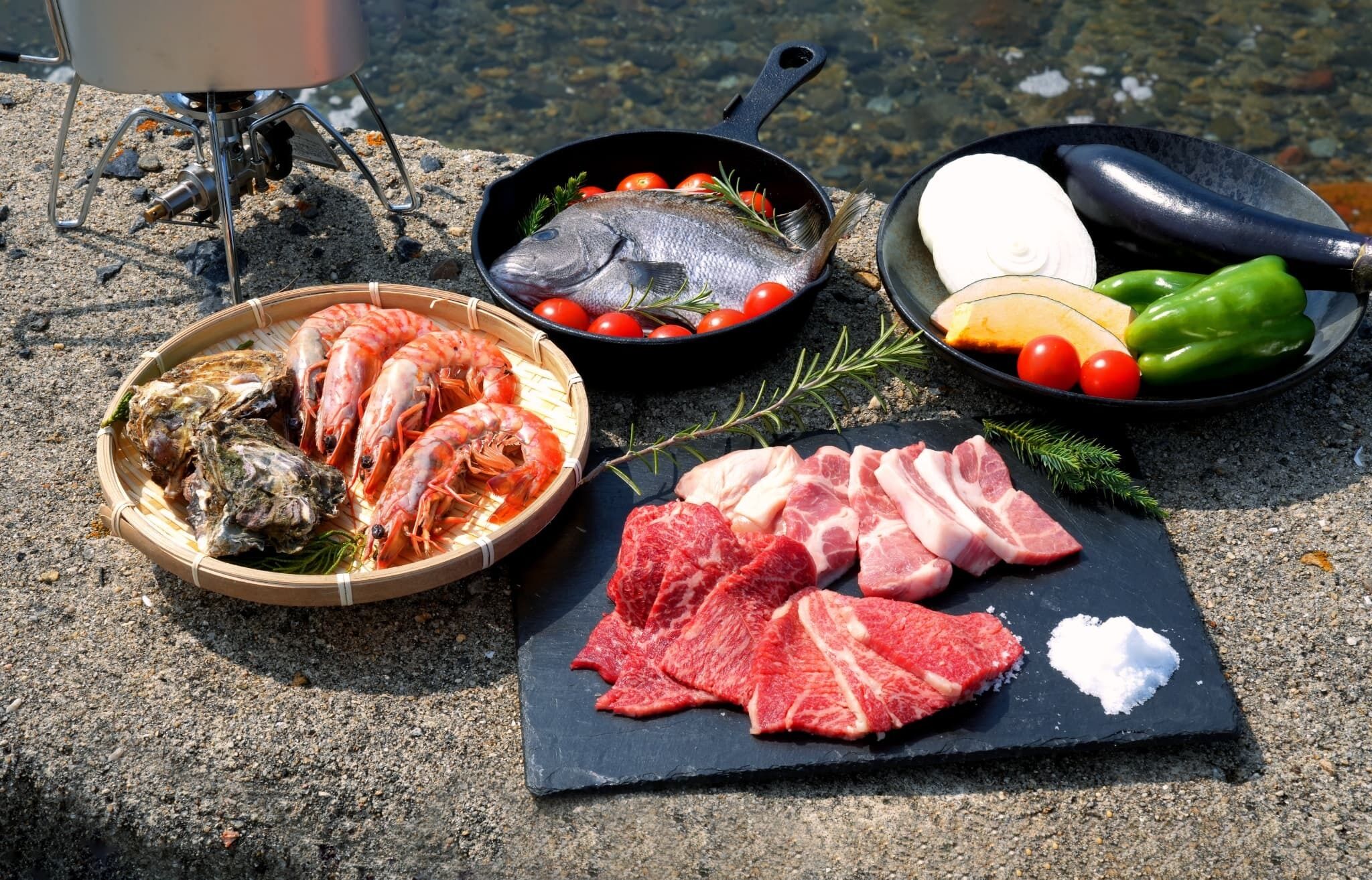 A variety of seafood and meats on a stone surface, including shrimp, fish, and steak, with cooking utensils nearby.