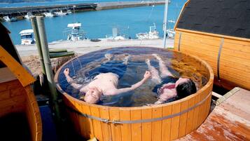 A wooden hot tub with two people inside, surrounded by a wooden deck and a view of the sea and boats.
