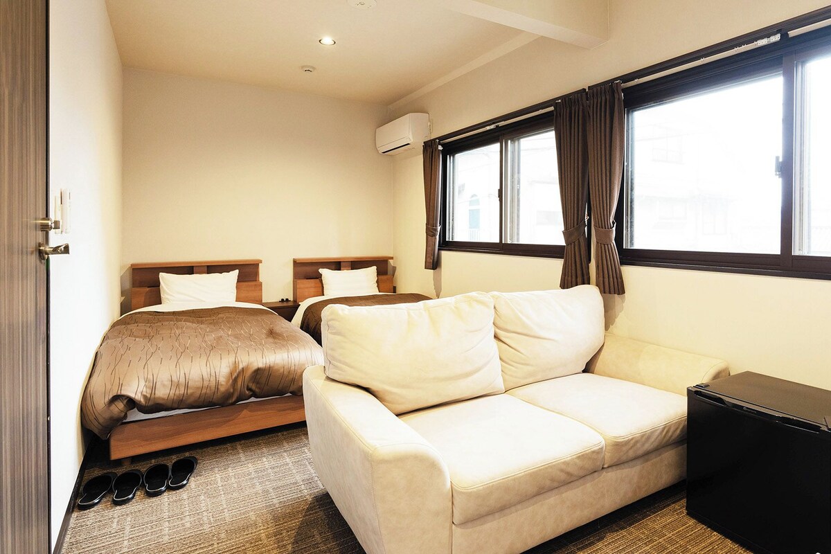 Habitación de hotel con dos camas, un sofá, una mesita, televisor y ventana con cortinas.