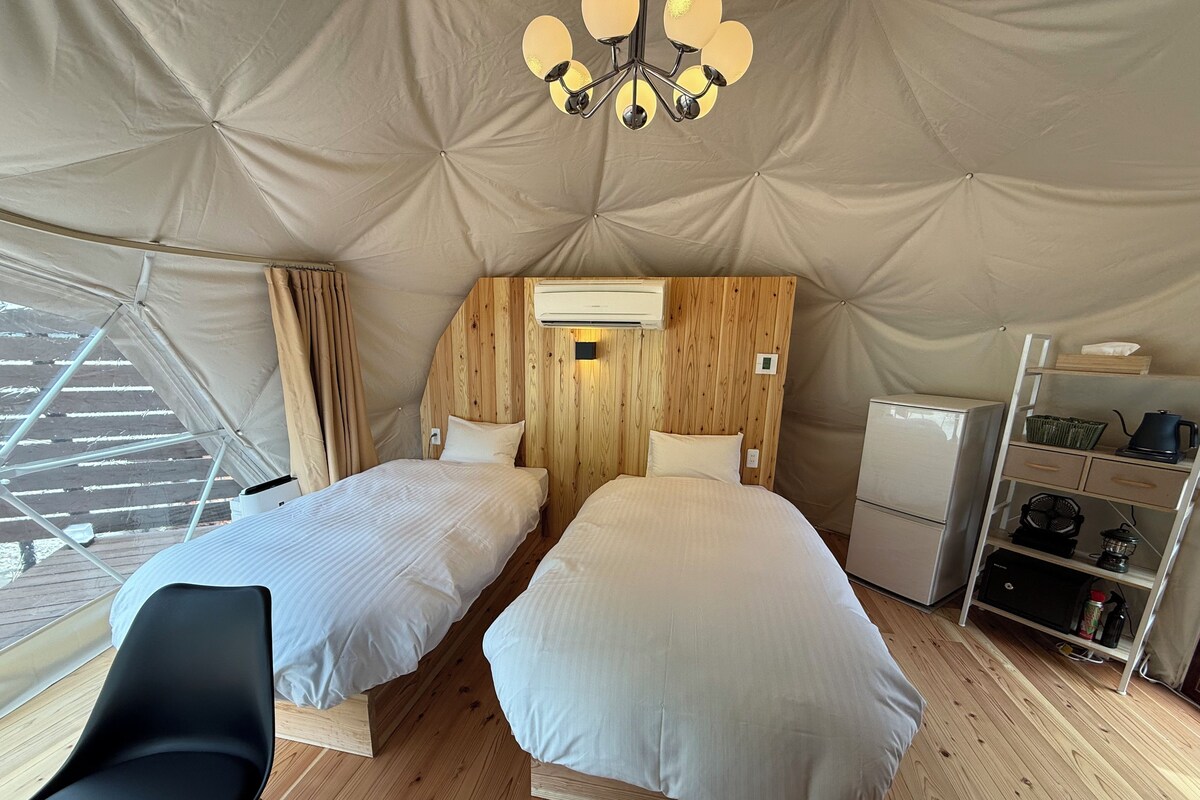 A tented accommodation with two beds, a wooden headboard, a small kitchenette, and a ceiling fan.