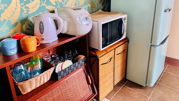 A kitchen with a microwave, kettle, and coffee maker on a shelf, a refrigerator, and wicker baskets with glassware. - Suite Terrace Ishigakijima (Ishigaki)