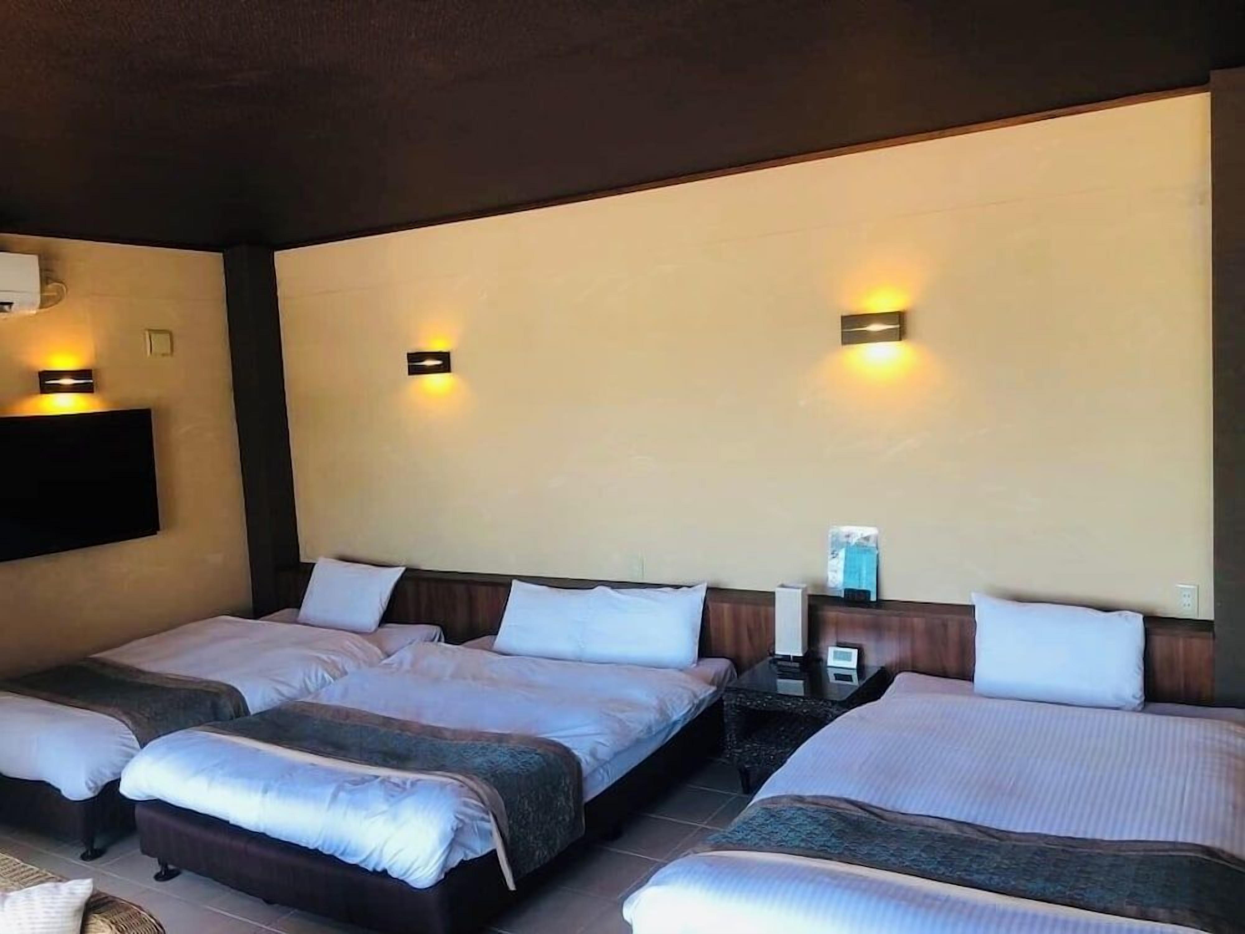 A hotel room with three beds arranged in a row, a flat-screen TV mounted on the wall, and two wall-mounted lamps.