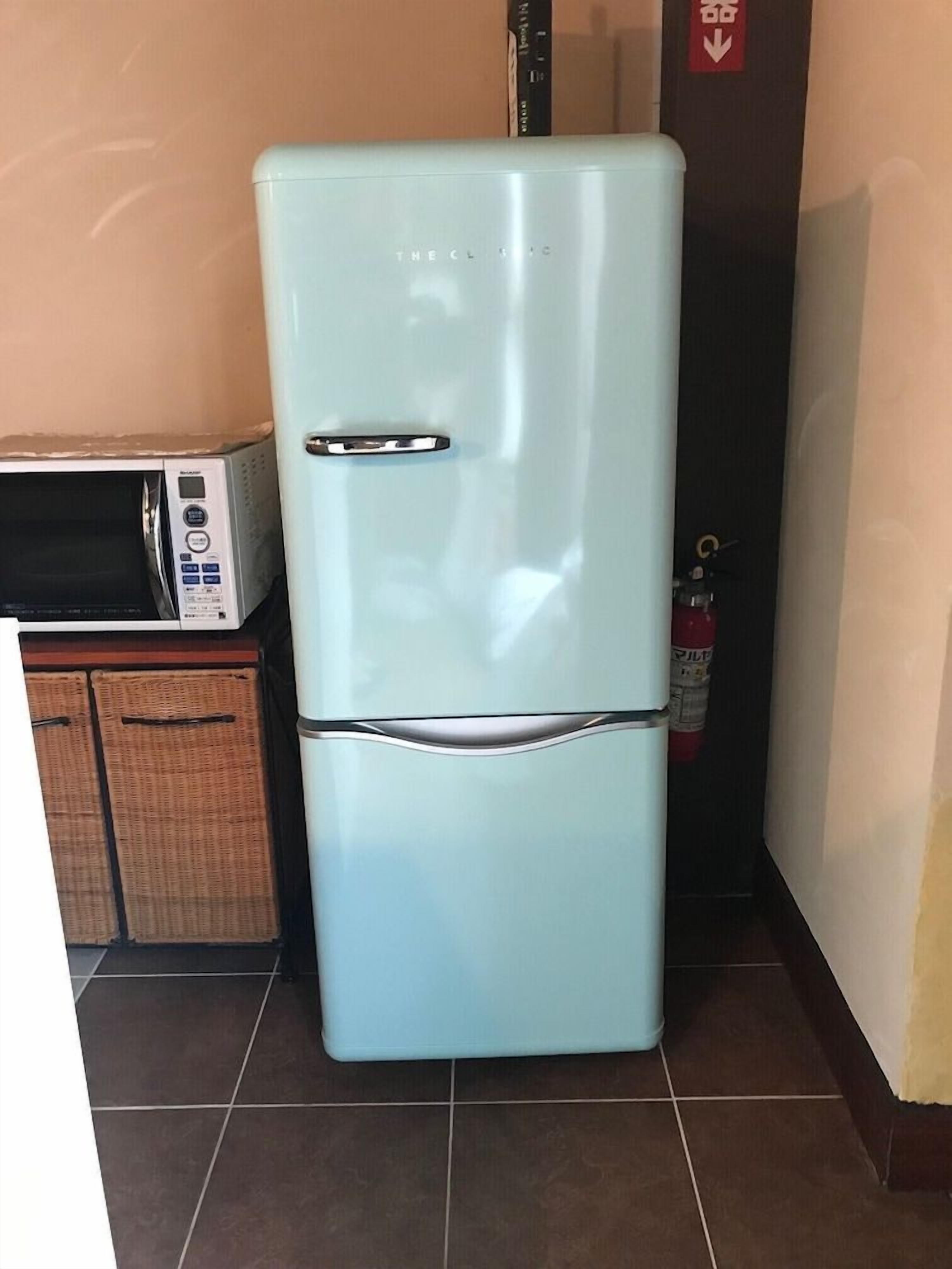 A light blue refrigerator in a kitchen with a microwave on top and a fire extinguisher in the background.