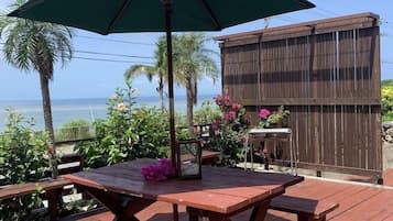 An outdoor patio with a wooden table and benches, a green umbrella, and a view of the ocean.