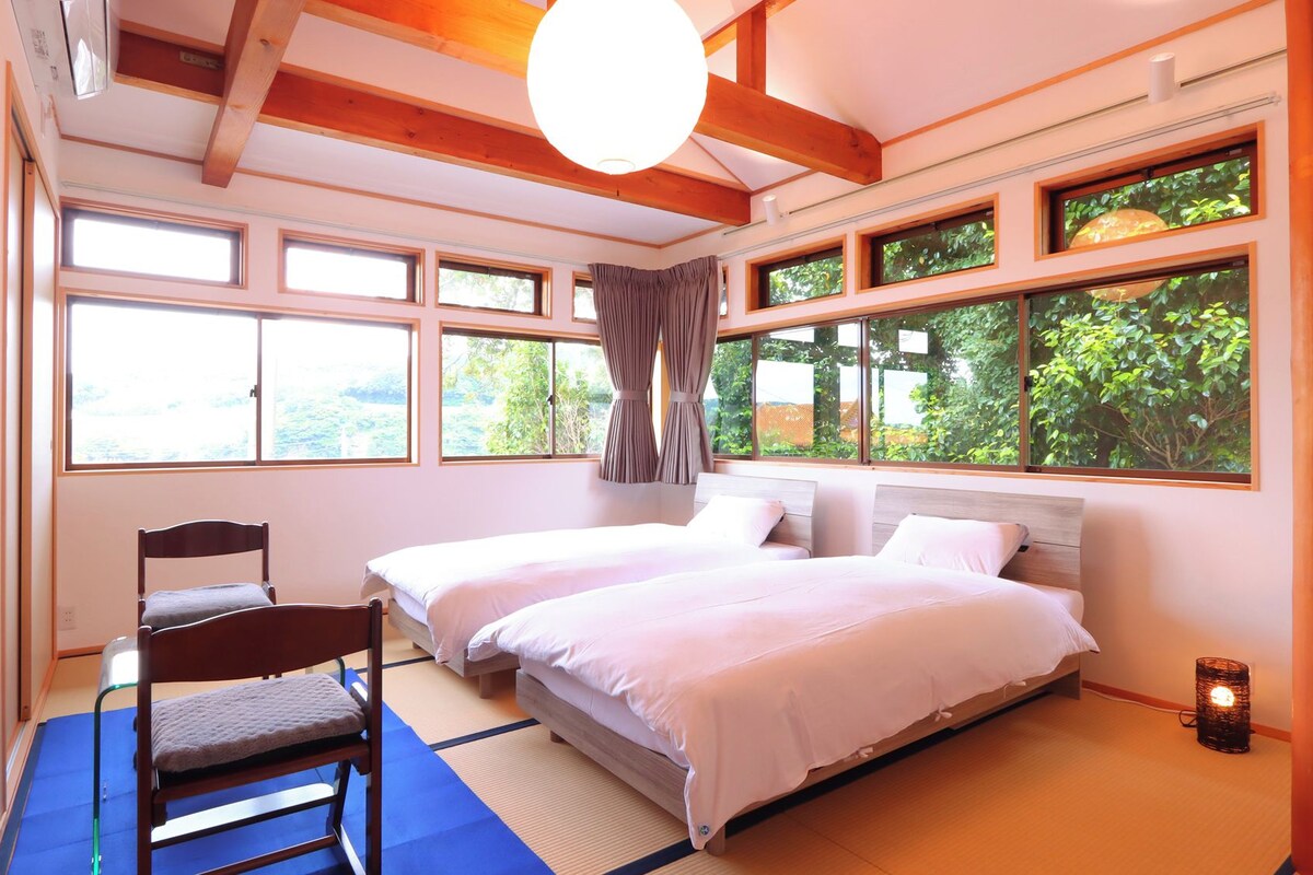 A room with two beds, a chair, tatami mats, large windows, and a view of greenery.