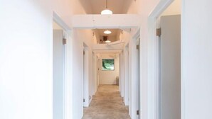 A narrow, well-lit hallway with white walls and doors on both sides, leading to a room with a window. - Hiruzen Shiogama Camping Village (Maniwa)